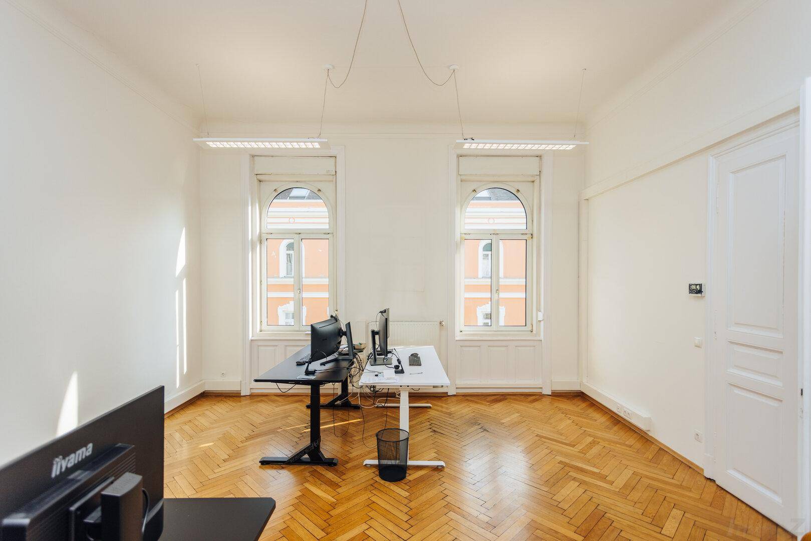 Helles Büro mit Fischgrätparkett, zwei großen Fenstern und einem Arbeitsplatz.