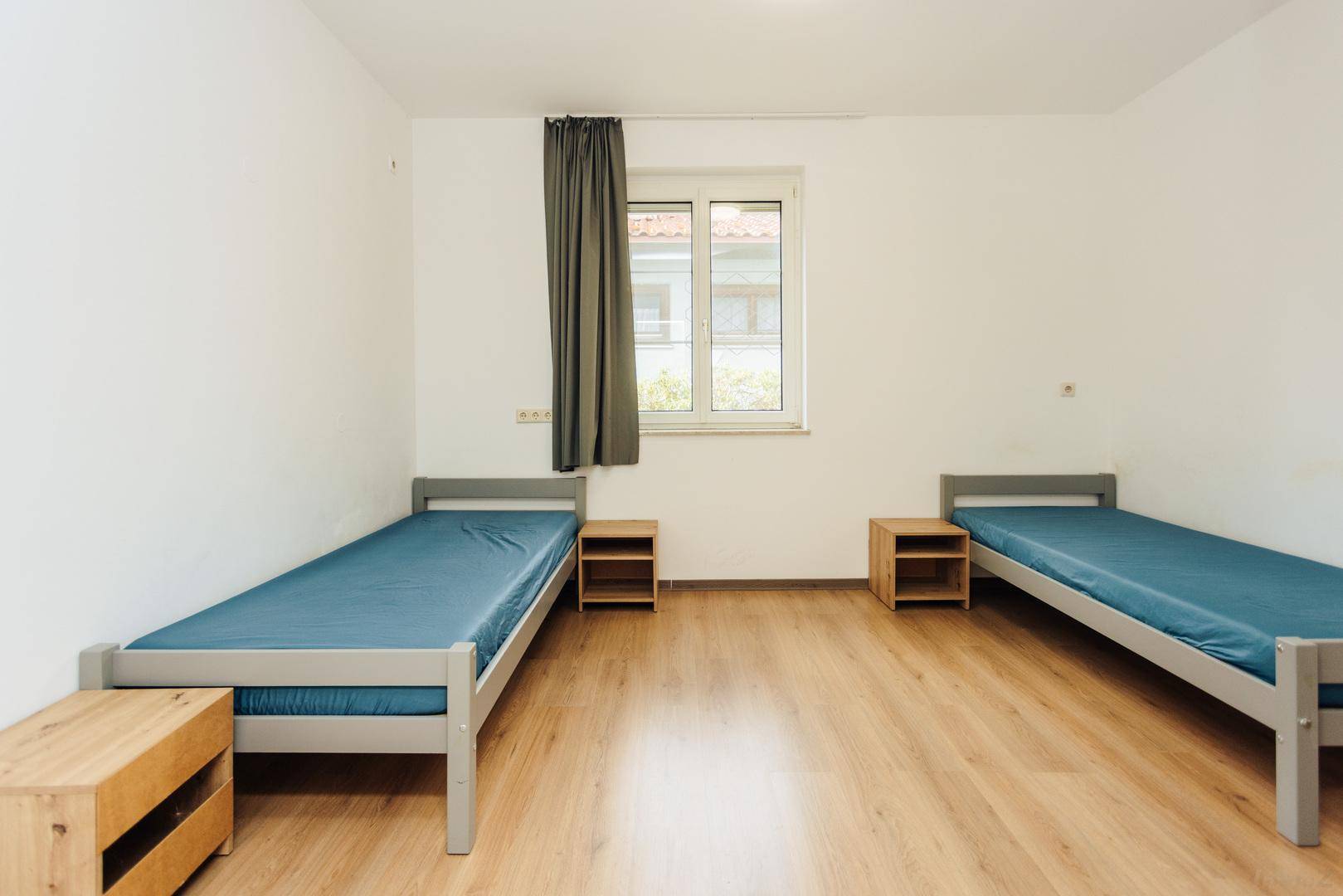 Einfaches Schlafzimmer mit zwei Einzelbetten, Holzmöbeln und einem Fenster.