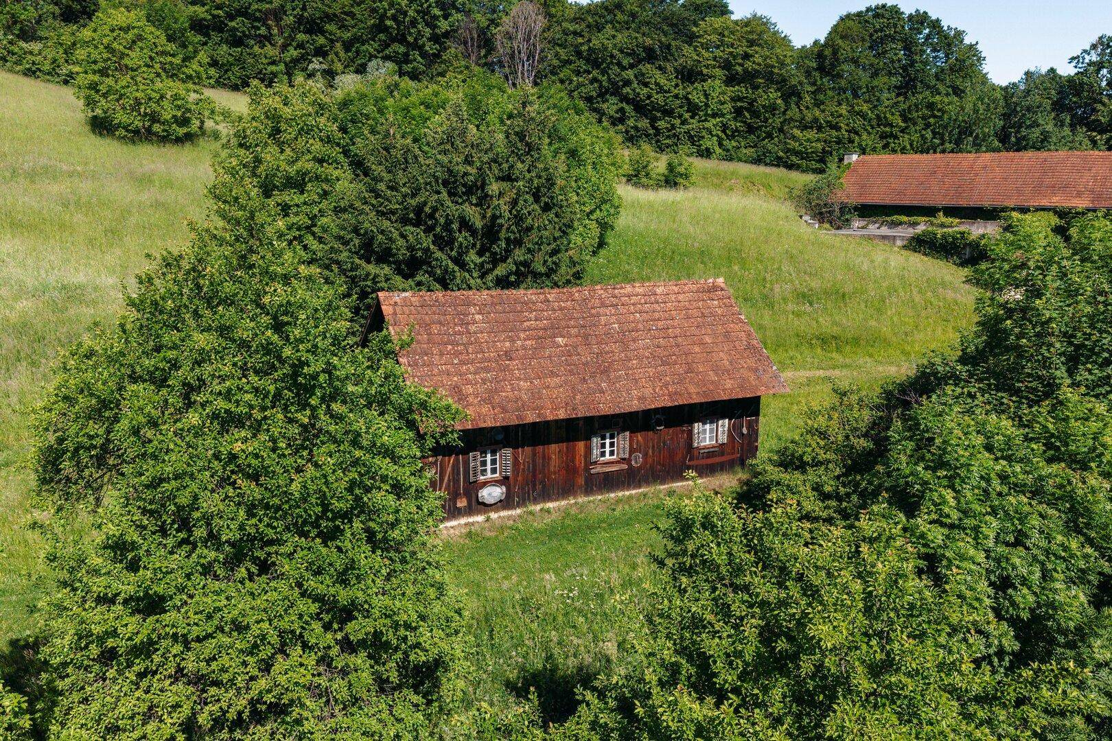 Kleineres Holzgebäude mit rotem Ziegeldach, eingebettet in die grüne Hügellandschaft und Bäume.
