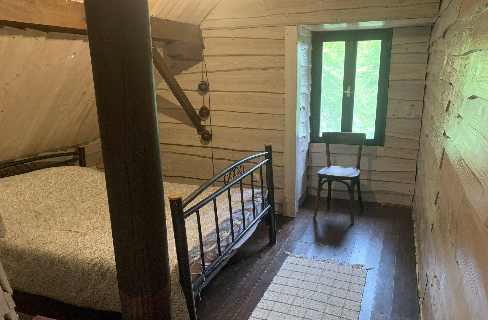 Einfaches Schlafzimmer mit Holzboden, Metallbett und einem Fenster mit Blick ins Grüne.