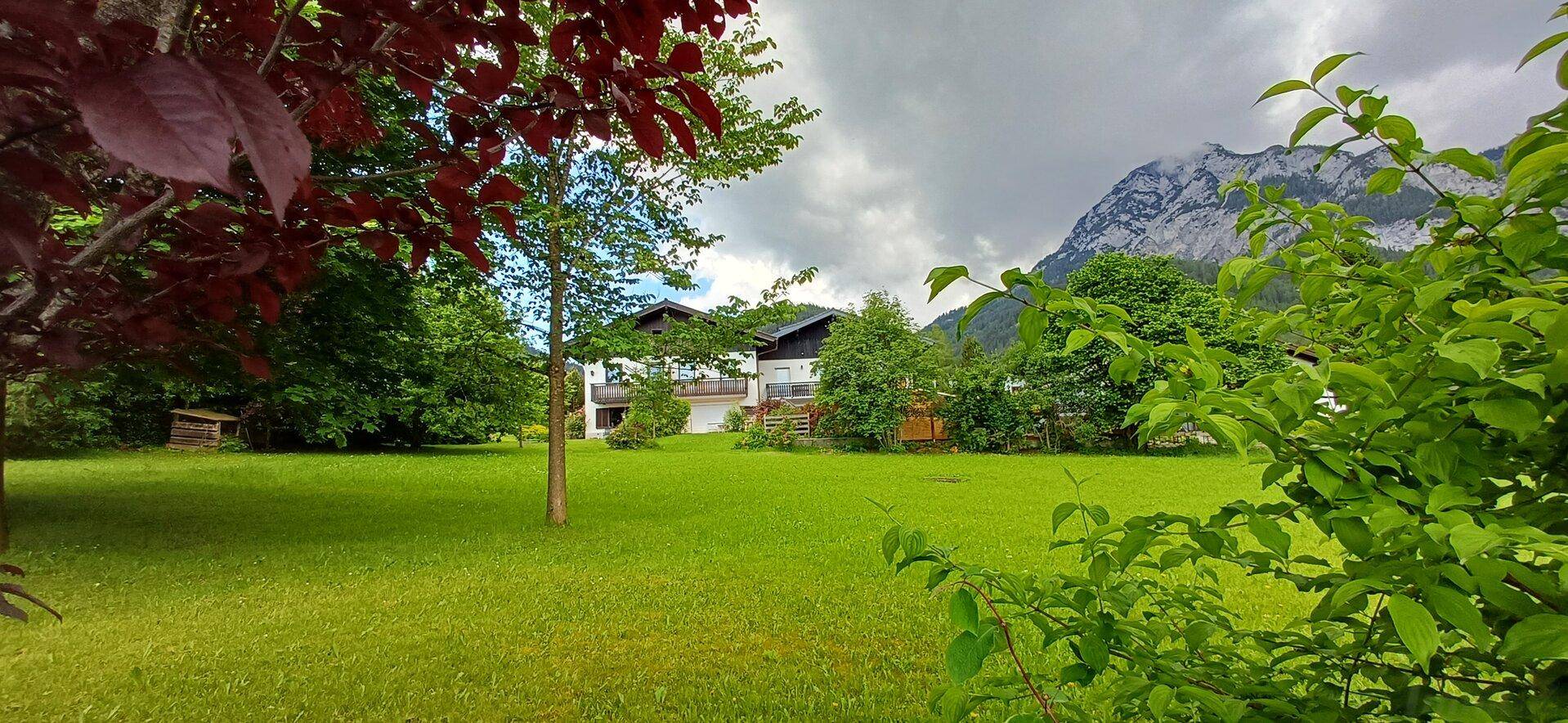Hausansicht von einem großen Rasen aus, umgeben von Bäumen und einer beeindruckenden Berglandschaft.