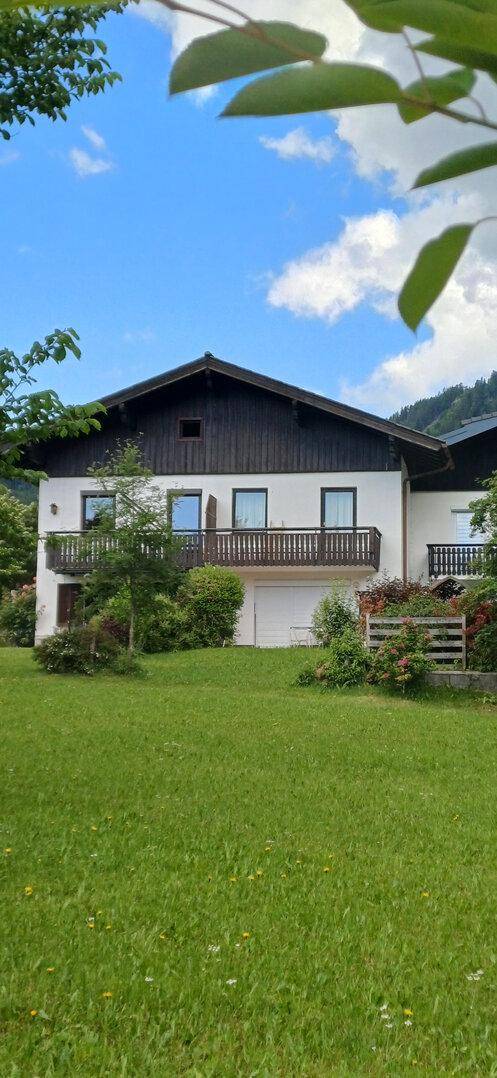Ansicht des Hauses mit Balkon und weitläufigem Rasen vor einer Bergkulisse unter blauem Himmel.