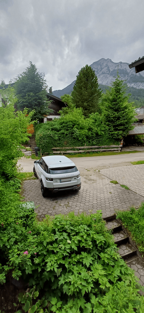 Gepflasterte Auffahrt mit einem Auto, umgeben von üppigem Grün und einer Bergkulisse.