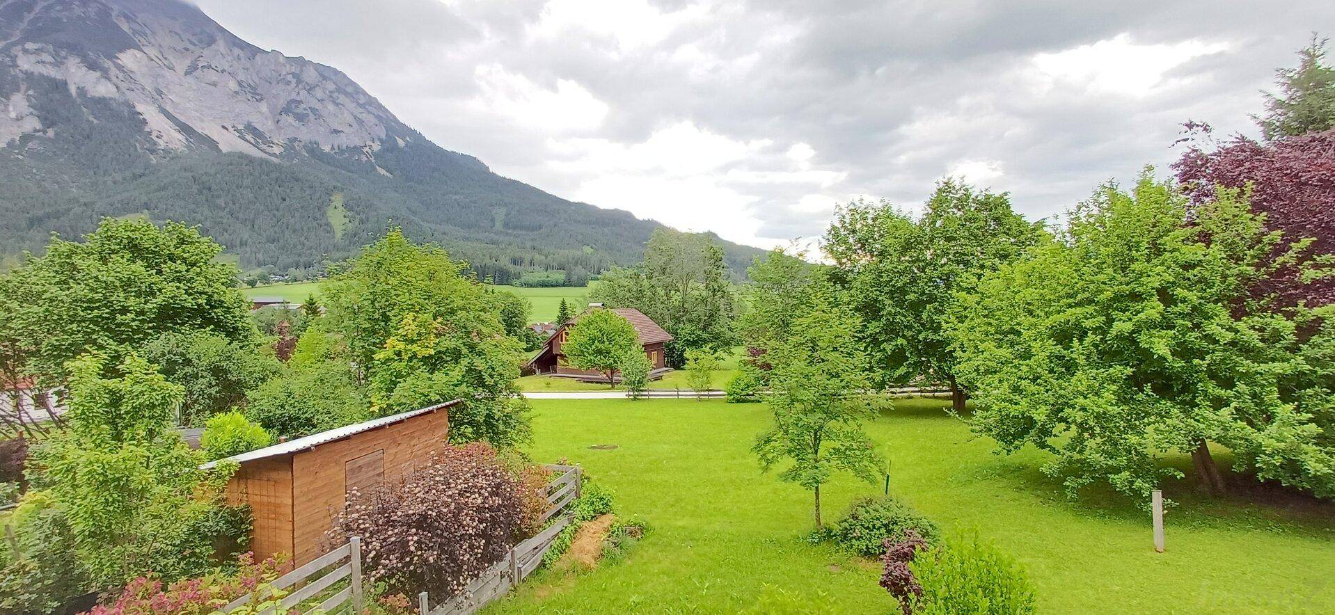 Weitläufige grüne Landschaft mit Bäumen, einem kleinen Schuppen und majestätischen Bergen im Hintergrund.