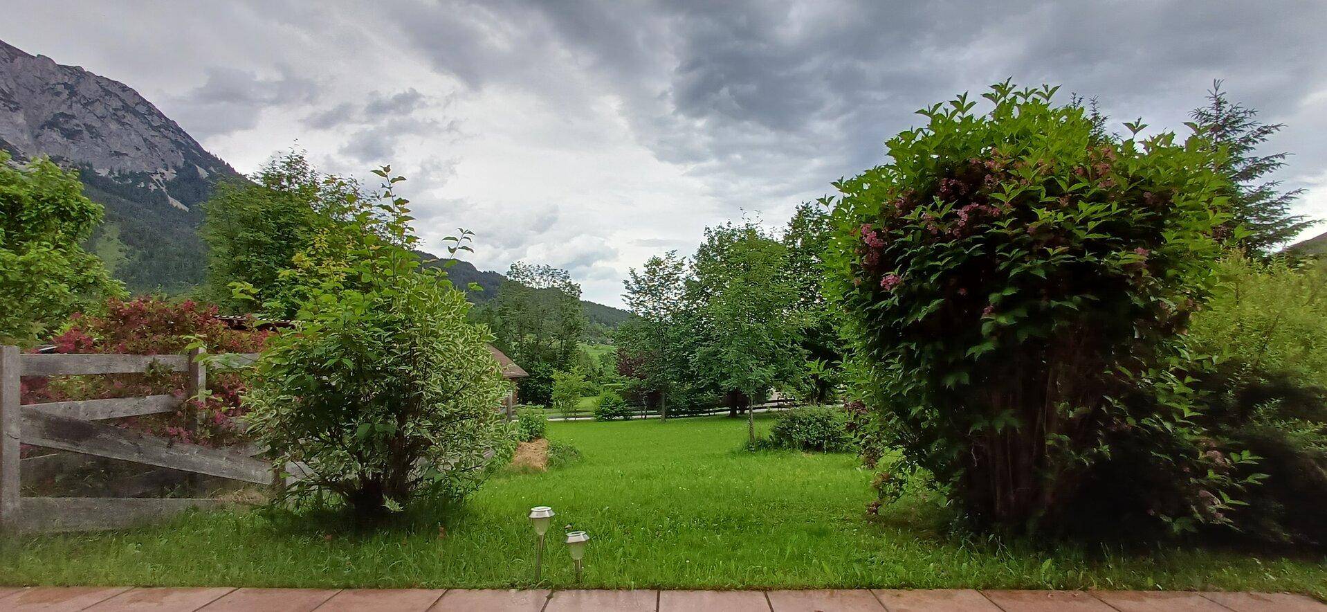 Blick von einer Terrasse in einen grünen Garten mit Sträuchern und Bergen im Hintergrund.