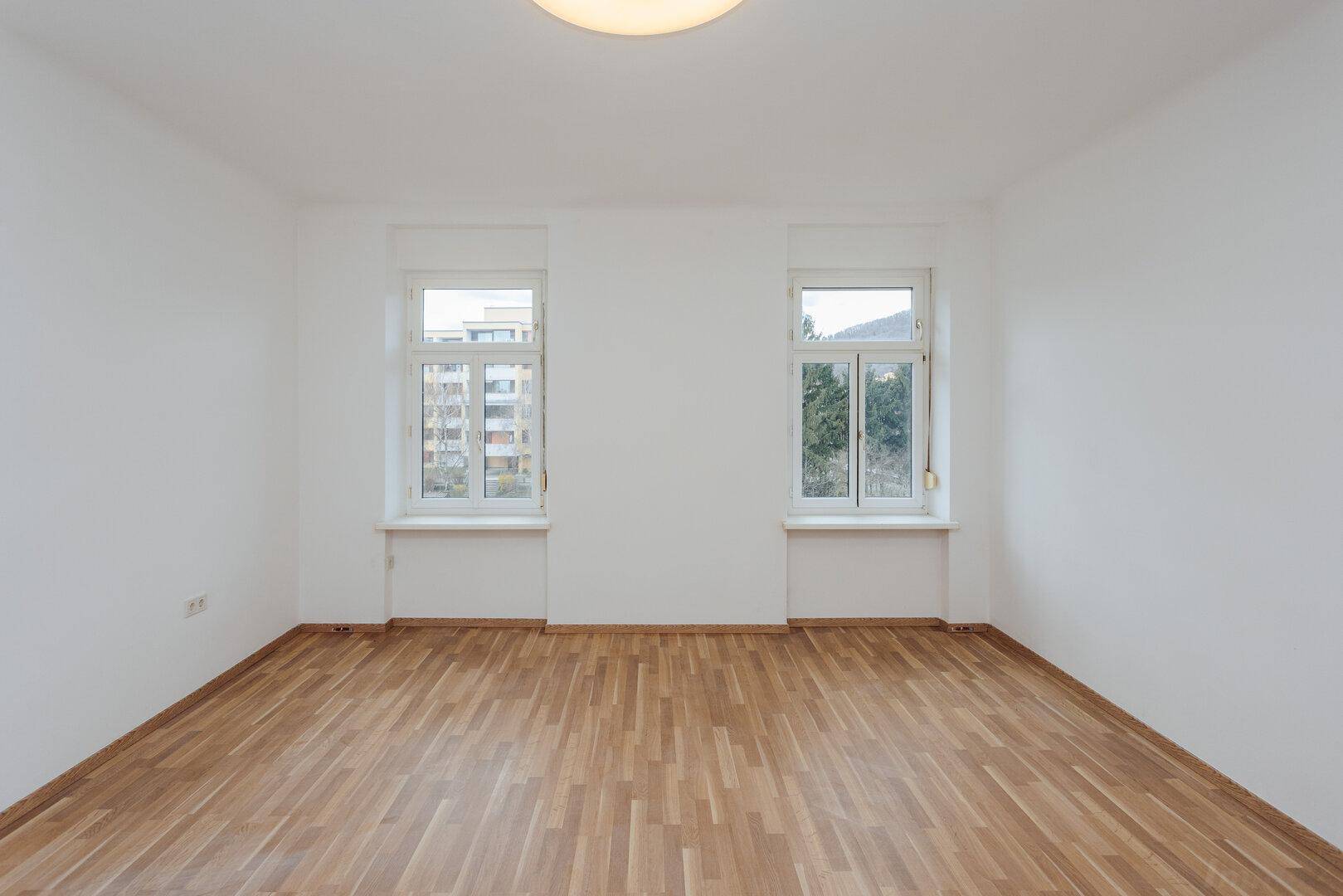 Geräumiges, leeres Zimmer mit Parkettboden und zwei Fenstern, die einen Blick nach draußen bieten.
