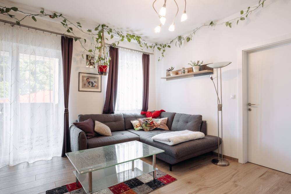 Gemütliches Wohnzimmer mit grauem Ecksofa, Holzboden und Zugang zu einem hellen Balkon.