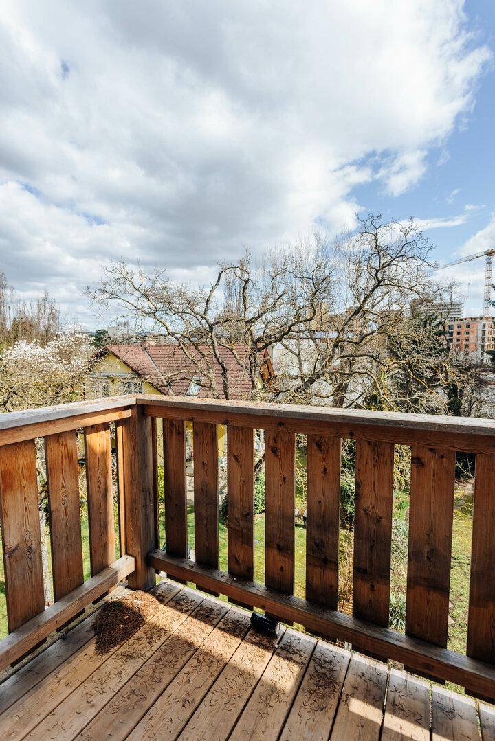 Rustikaler Holzbalkon mit Geländer und Blick auf Bäume und umliegende Gebäude unter blauem Himmel.