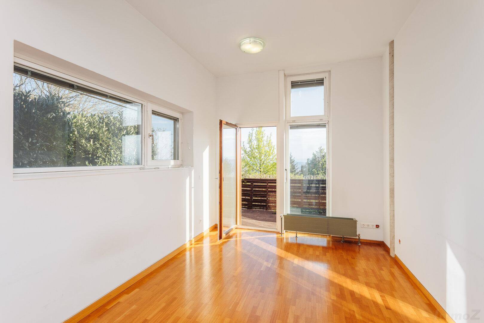 Leeres Zimmer mit Holzboden, großen Fenstern und Zugang zu einem Außenbereich.