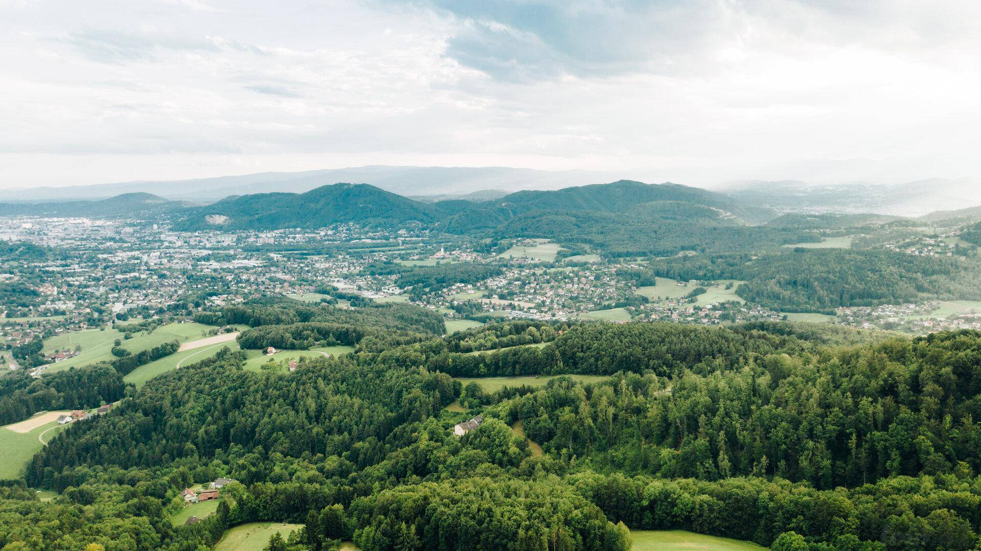 Weitläufige Luftaufnahme einer grünen Hügellandschaft mit Wäldern und Siedlungen im Hintergrund.