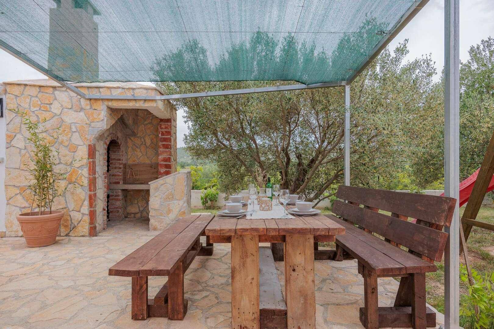 Rustikaler Grillbereich im Freien mit gemauertem Grill und Holztischgarnitur unter einem Sonnenschutz.