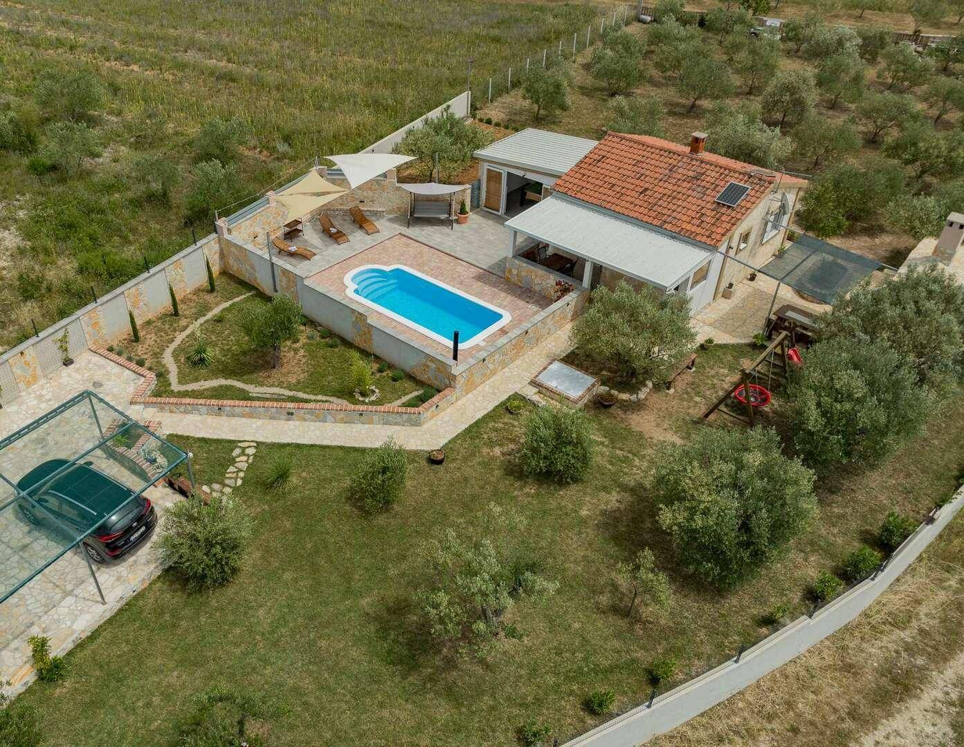 Luftaufnahme eines Hauses mit Pool, Gartenbereich und überdachtem Parkplatz in ländlicher Umgebung.