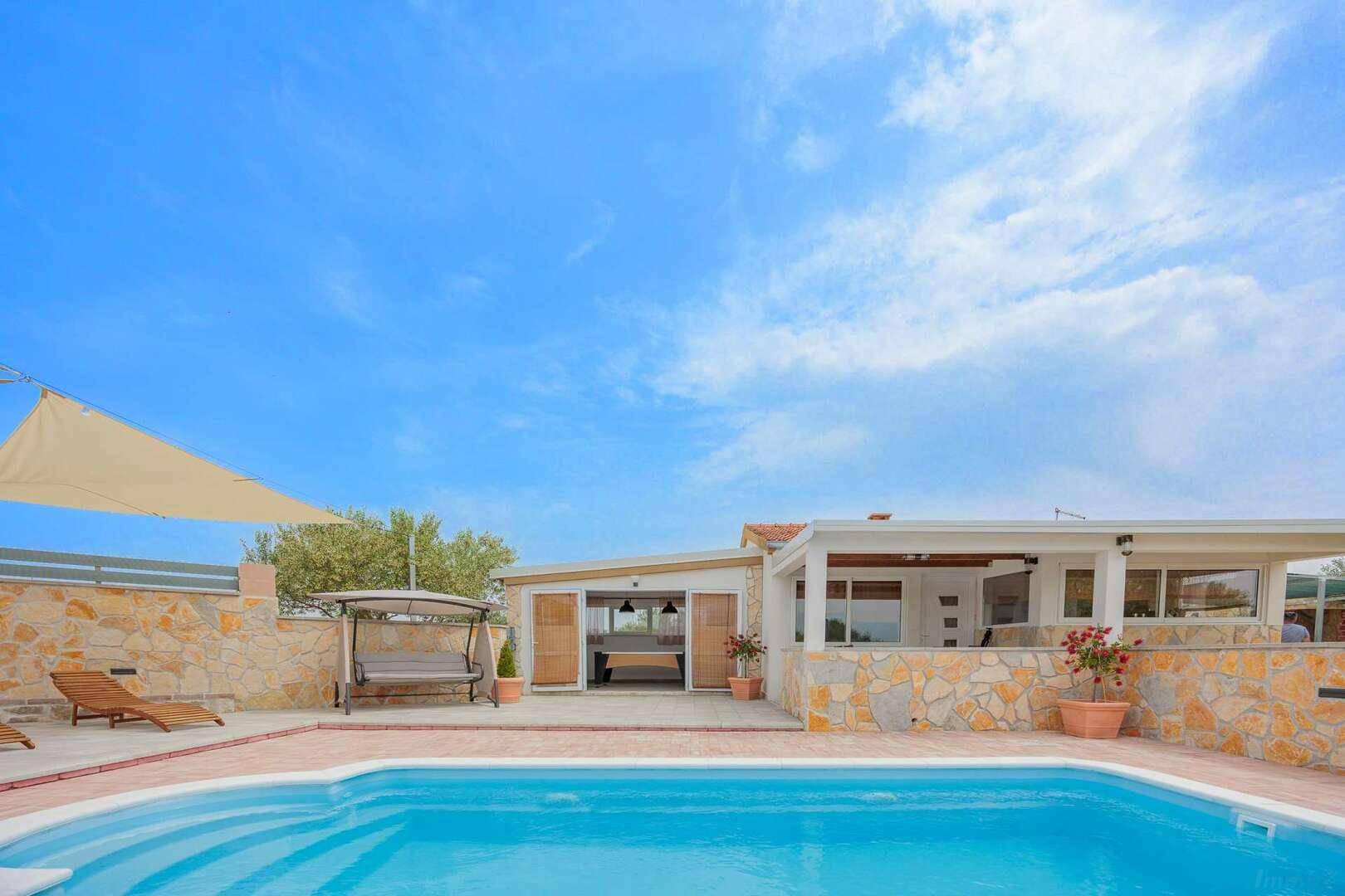 Blick auf den Poolbereich mit Liegestühlen, Sonnensegeln und einer überdachten Terrasse am Haus.