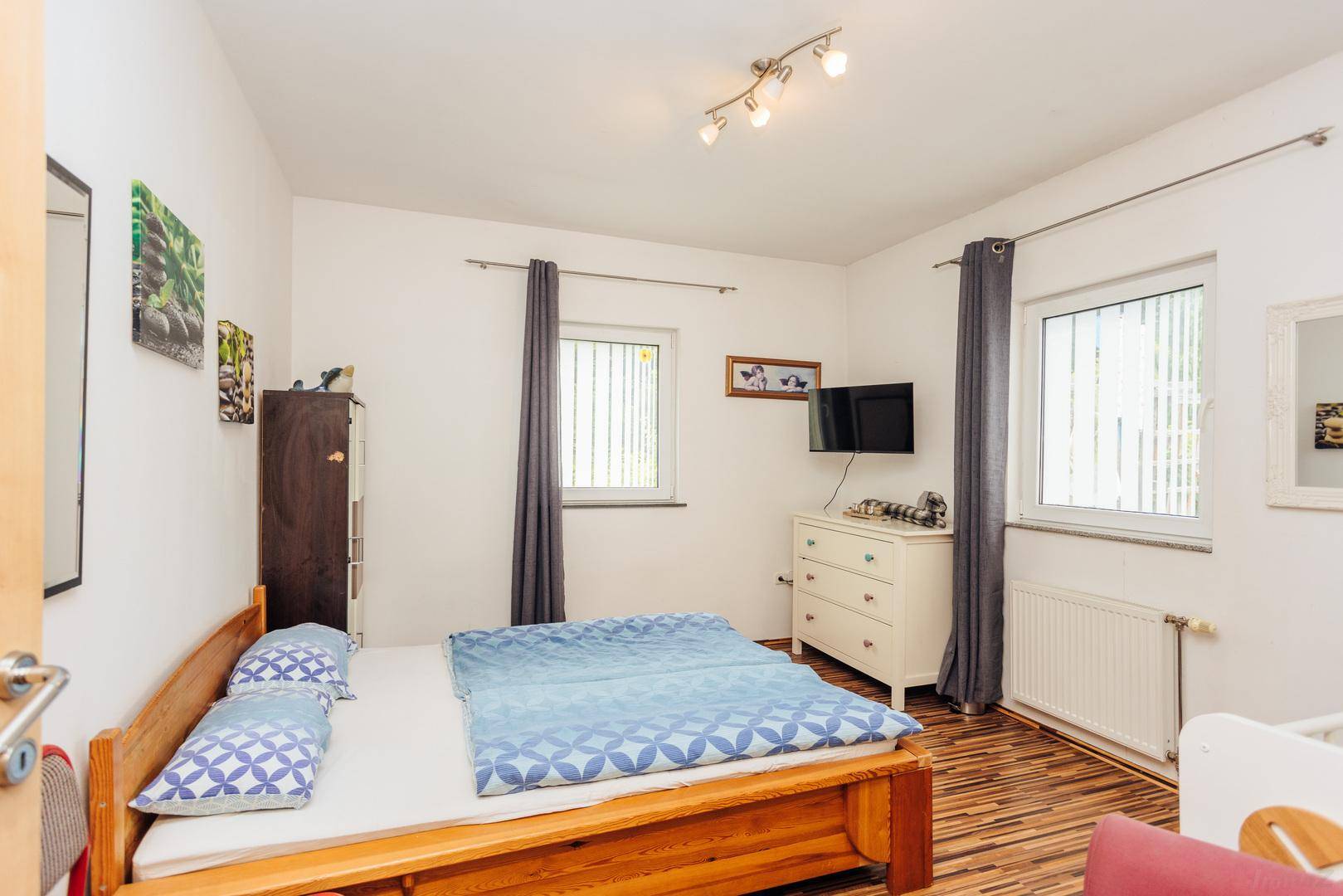 Schlafzimmer mit Holzbett, Kommode und zwei Fenstern für natürliches Licht.