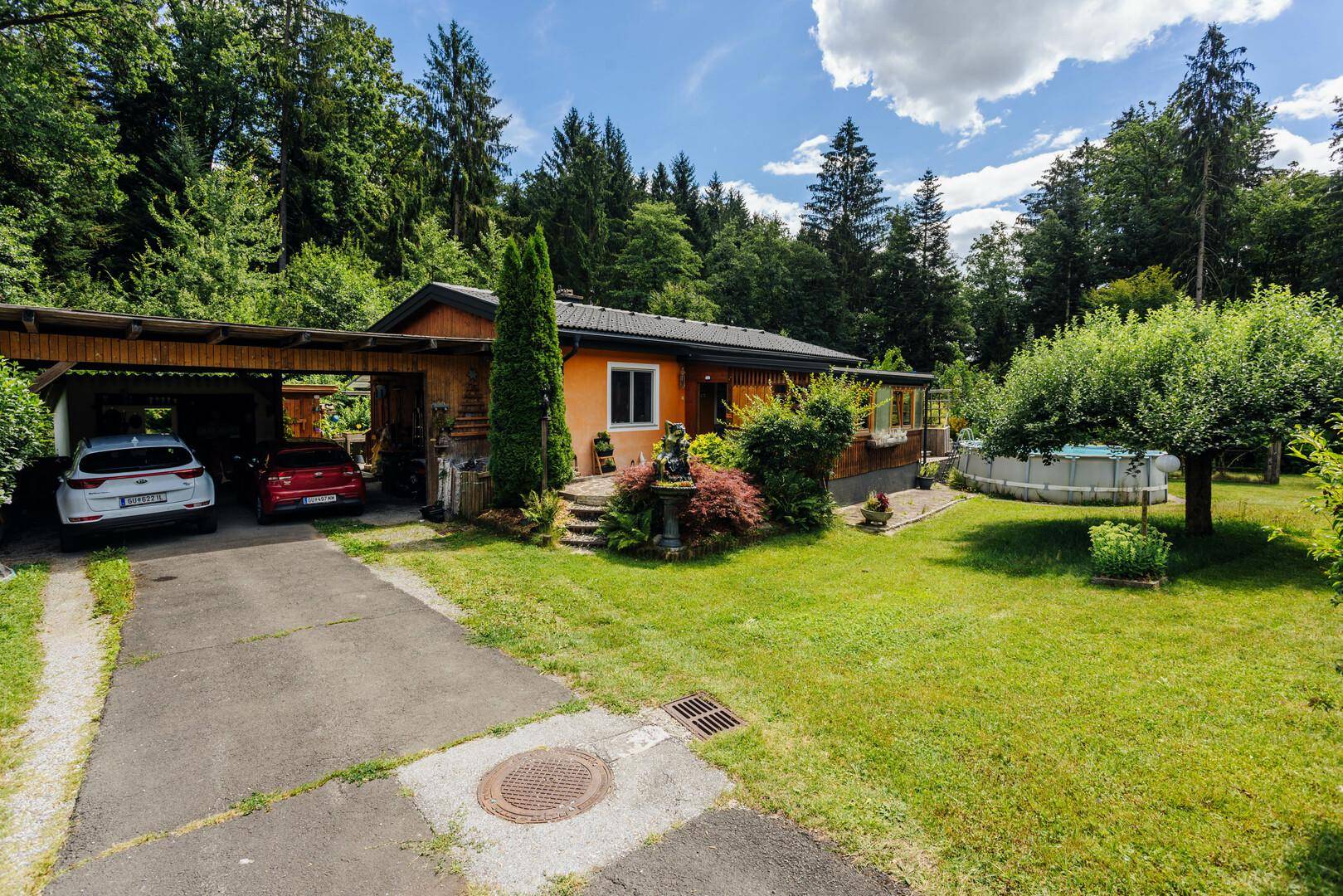 Hausansicht mit Carport für zwei Fahrzeuge, umgeben von einem grünen Garten und Wald.