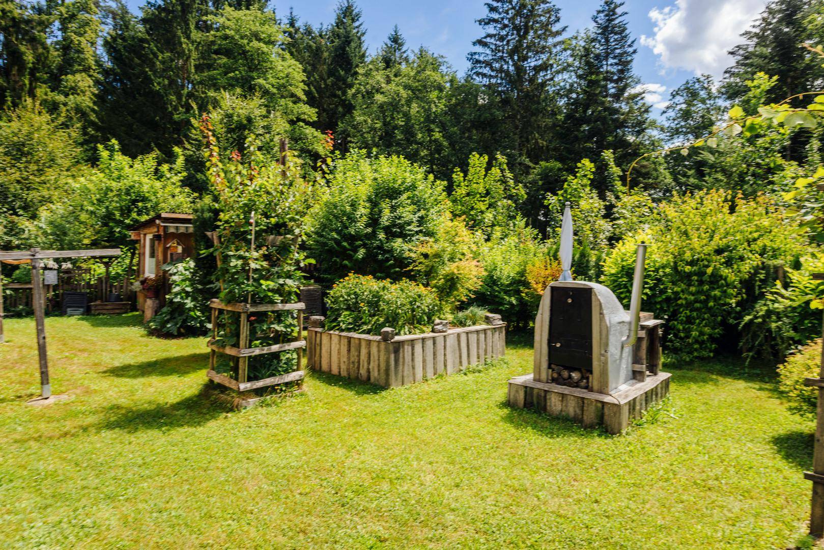 Gartenbereich mit Hochbeeten, einem Außenofen und üppiger Bepflanzung.