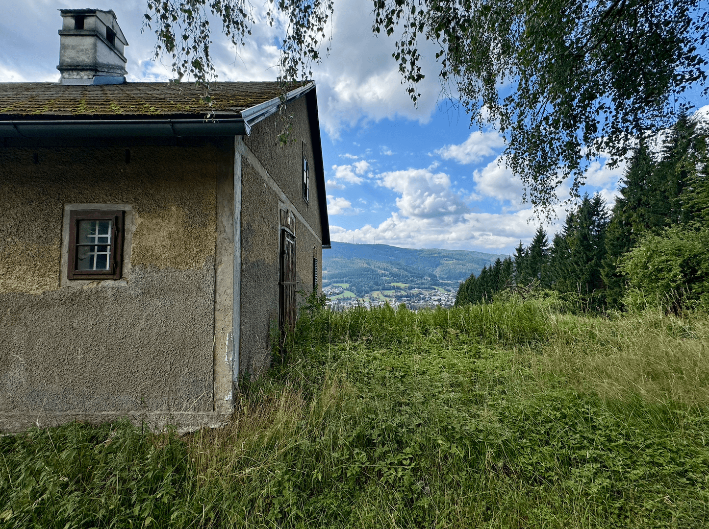 Seitenansicht des alten Hauses mit Schornstein und weitem Blick über die bewaldete Hügellandschaft.