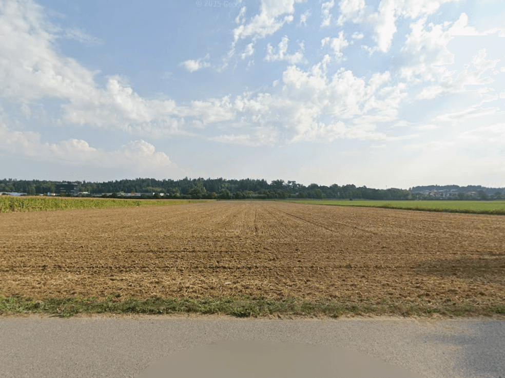 Weites Feld mit Ackerland unter blauem Himmel, umgeben von Bäumen und ländlicher Bebauung.