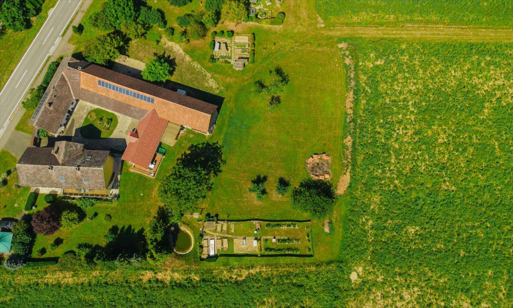 Luftaufnahme des Anwesens mit Gebäuden, Gartenbereichen und umliegenden Feldern unter blauem Himmel.