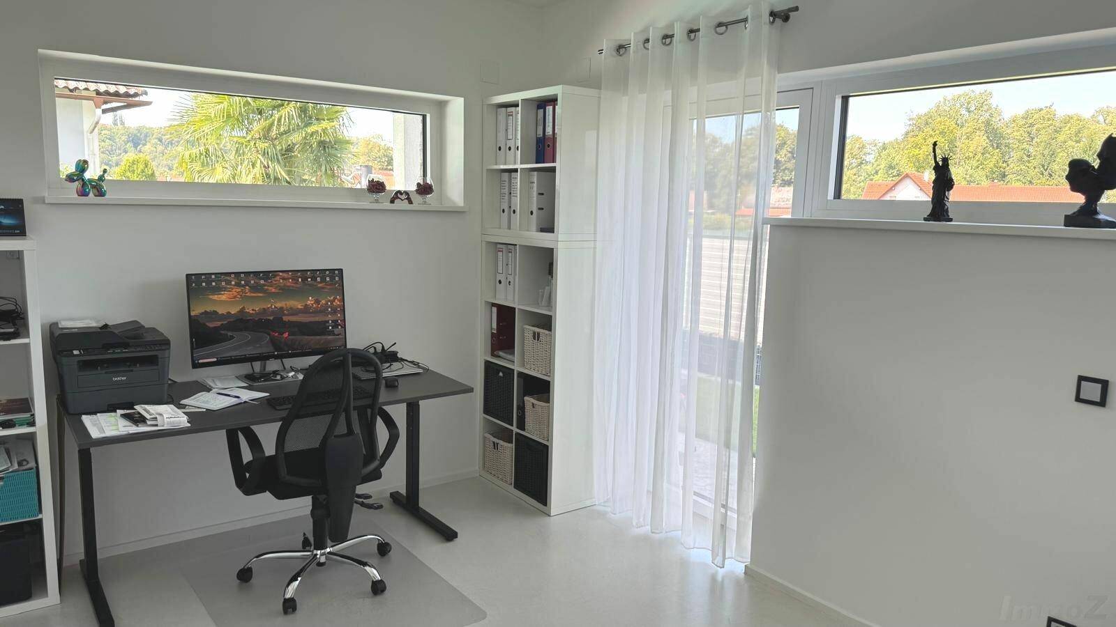 Helles Arbeitszimmer mit Schreibtisch, Regalen und großen Fenstern mit Blick ins Grüne.