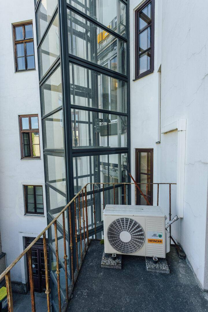 Außenansicht eines Gebäudes mit einem modernen Glasaufzug und einer Klimaanlage auf einem kleinen Balkon.