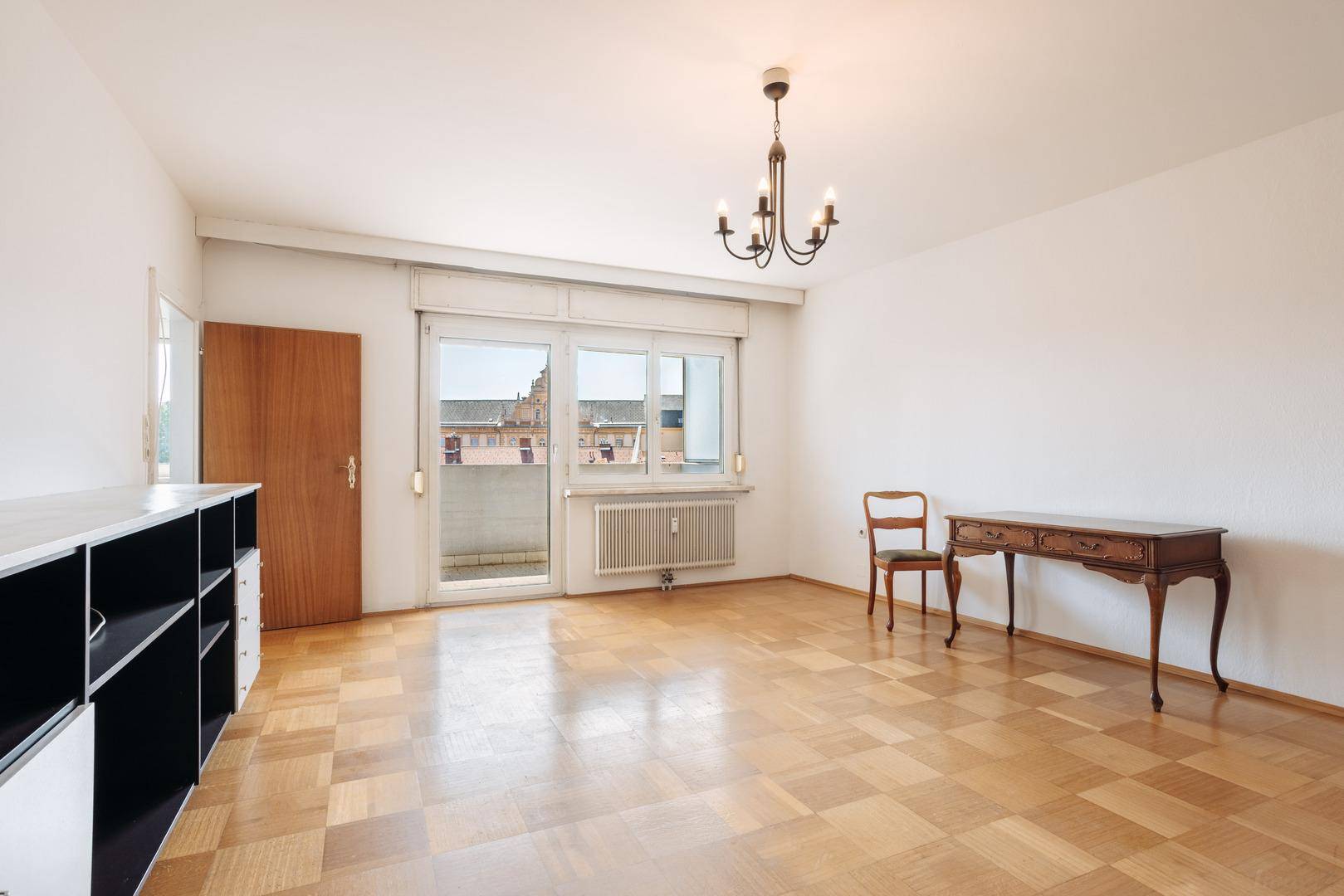 Helles Wohnzimmer mit Parkettboden, Zugang zum Balkon und einfacher Einrichtung.