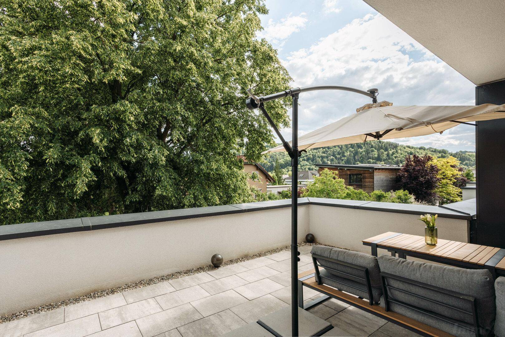 Große, geflieste Terrasse mit Sitzgelegenheiten, Sonnenschirm und Blick ins Grüne.