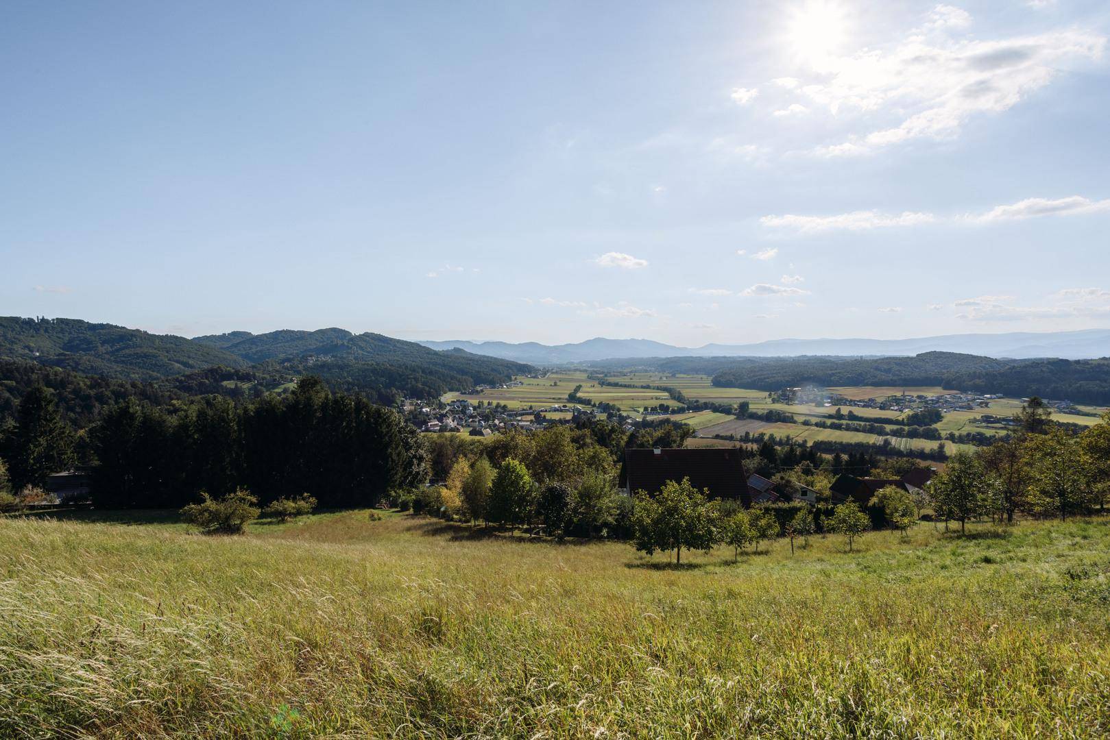 Weitläufiger Blick über eine grüne Hügellandschaft mit Feldern und einem Dorf in der Ferne unter blauem Himmel.