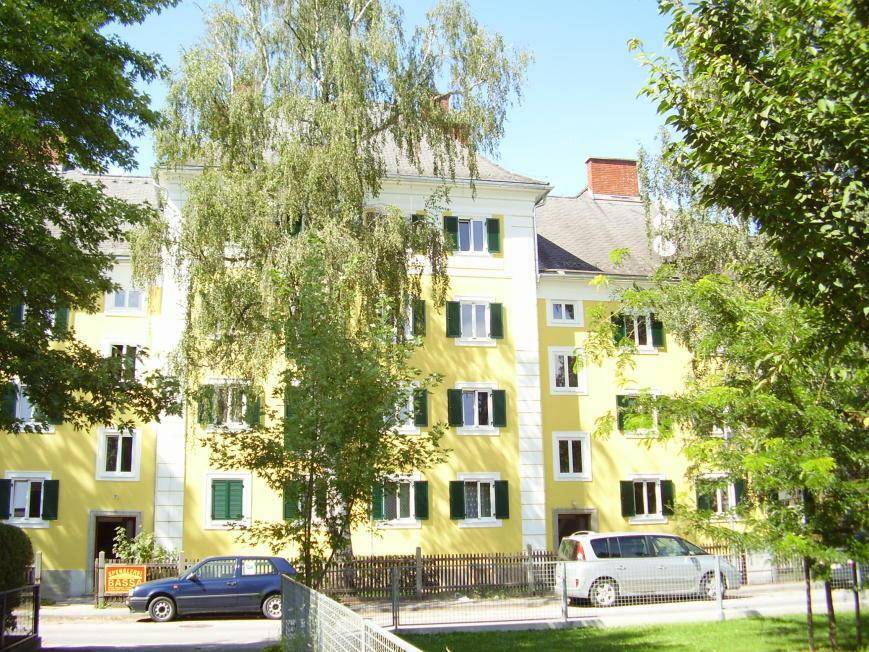 Gelbes Wohngebäude mit grünen Fensterläden und Bäumen im Vordergrund, geparkte Autos auf der Straße.