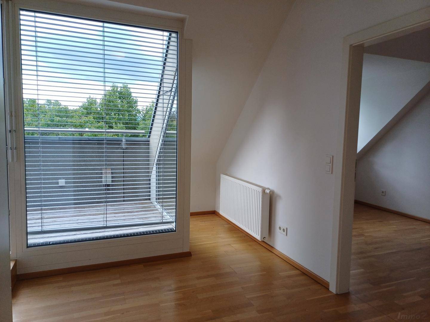 Heller Raum im Dachgeschoss mit Holzboden, Heizkörper und großem Fenster mit Zugang zu einem Außenbereich.