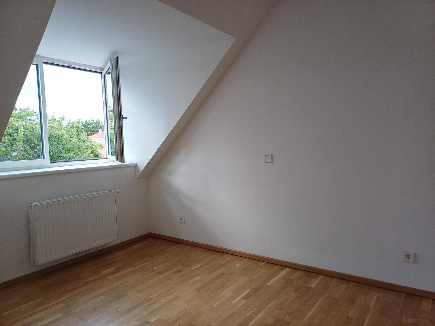 Geräumiges Zimmer im Dachgeschoss mit Holzboden, Heizkörper und Fenster mit Blick ins Grüne.