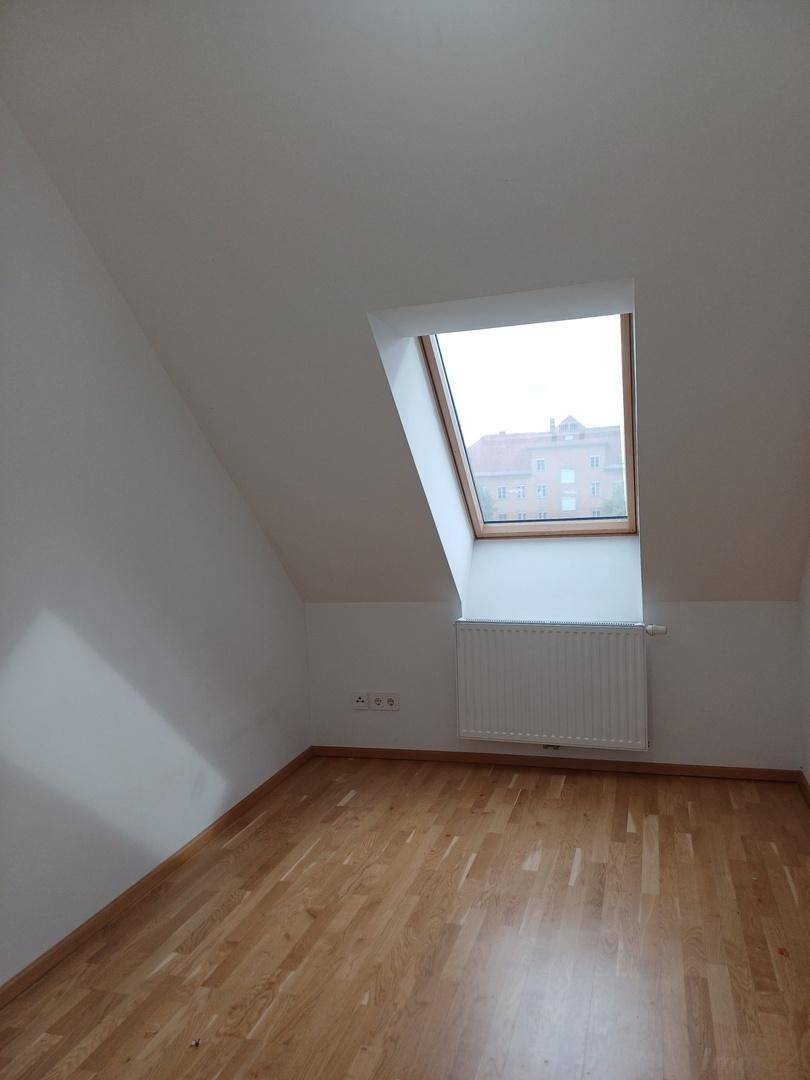 Helles Zimmer im Dachgeschoss mit Holzboden, Heizkörper und einem Dachfenster für natürliches Licht.