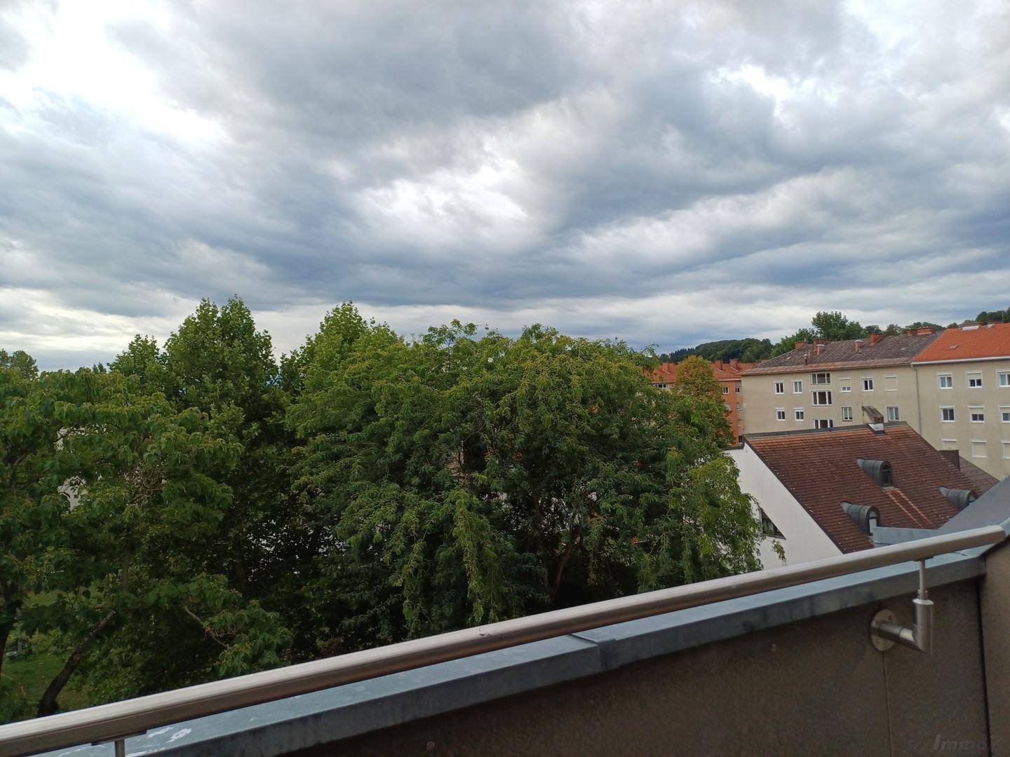 Aussicht von einem Balkon mit Blick auf grüne Bäume und umliegende Wohngebäude unter bewölktem Himmel.