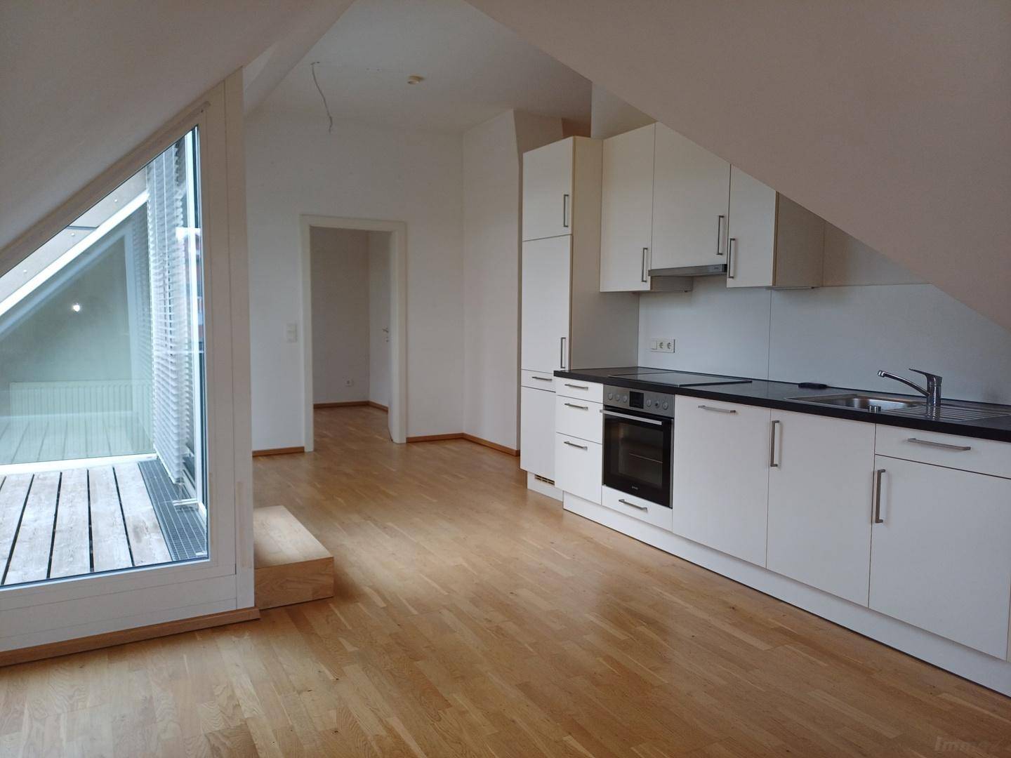Heller Wohnbereich mit integrierter Küche, Holzboden und großem Fenster mit Zugang zu einem Außenbereich.