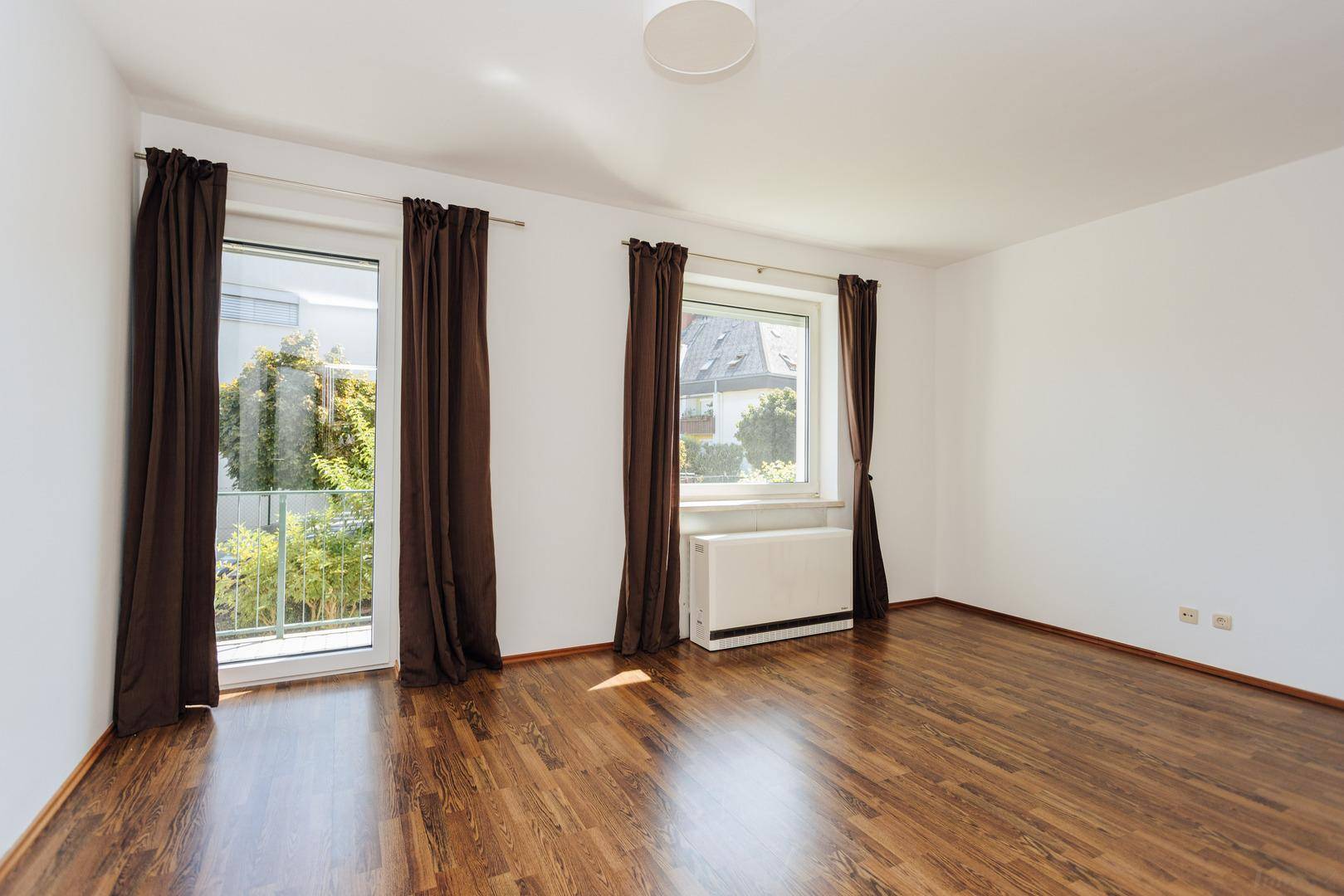 Leeres Zimmer mit Holzboden, großen Fenstern und Zugang zu einem Balkon.