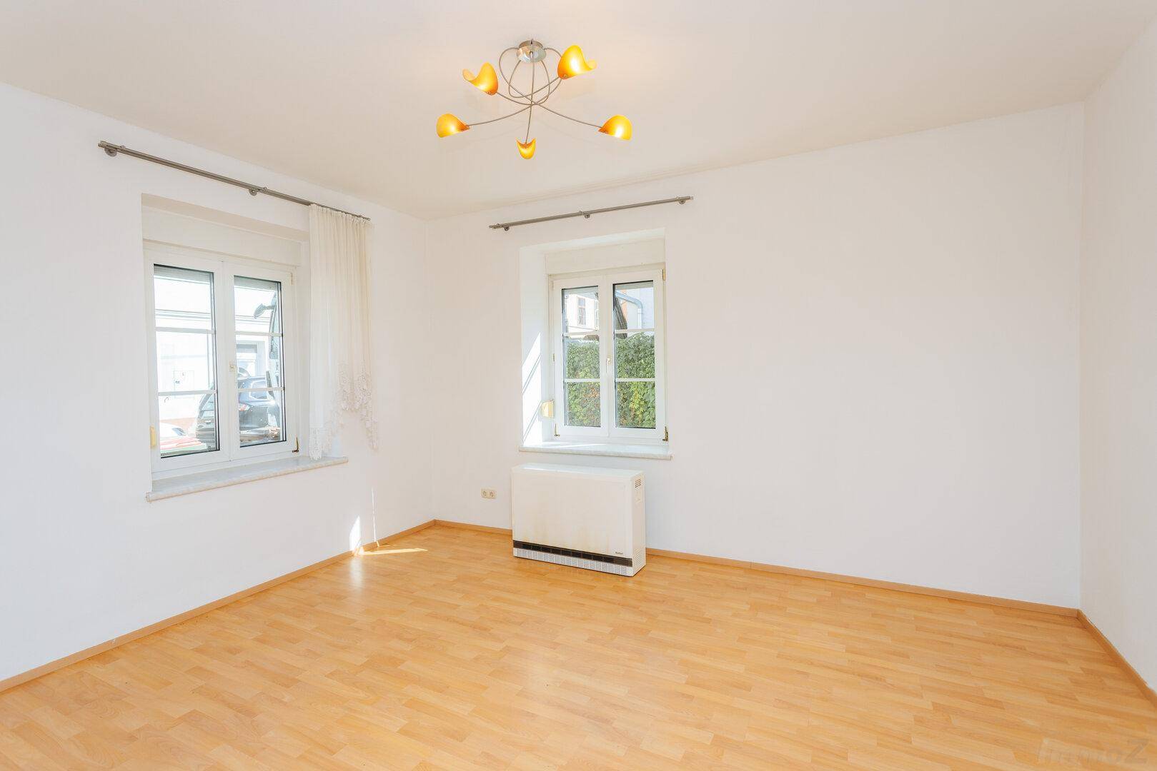 Leeres Wohnzimmer mit hellem Holzboden, weißen Wänden und zwei Fenstern für viel Tageslicht.