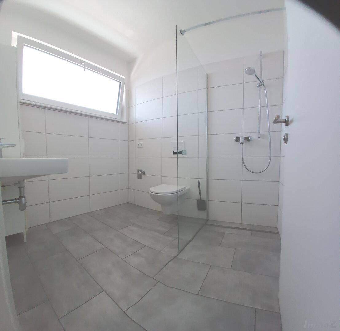 Modernes Badezimmer mit ebenerdiger Dusche, wandhängendem WC und Fenster für Tageslicht, komplett gefliest.