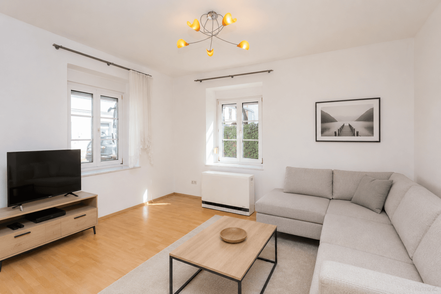 Helles Wohnzimmer mit modernem Sofa, Holzboden und zwei Fenstern, die viel Tageslicht hereinlassen.
