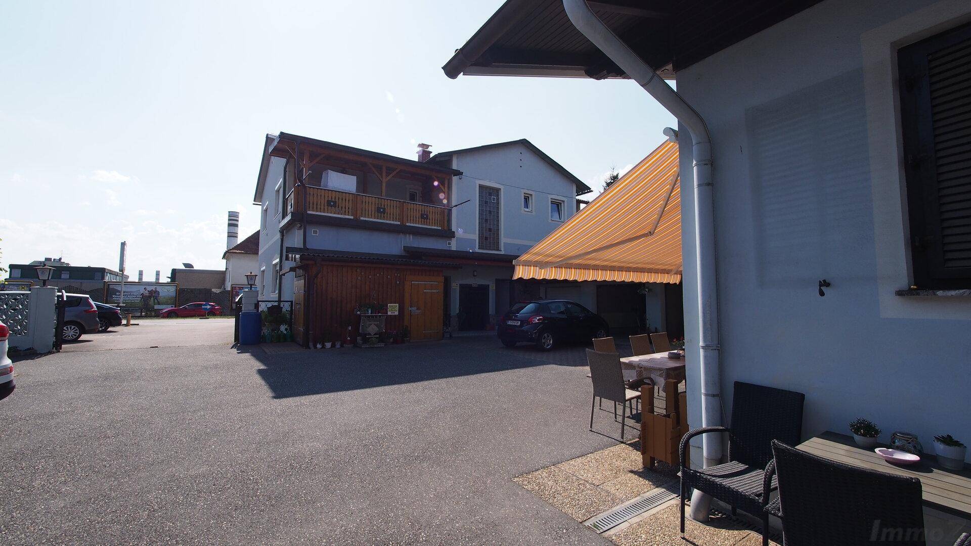 Außenbereich mit gepflastertem Parkplatz, Sitzgelegenheiten und einem Gebäude mit Balkon und Markise.