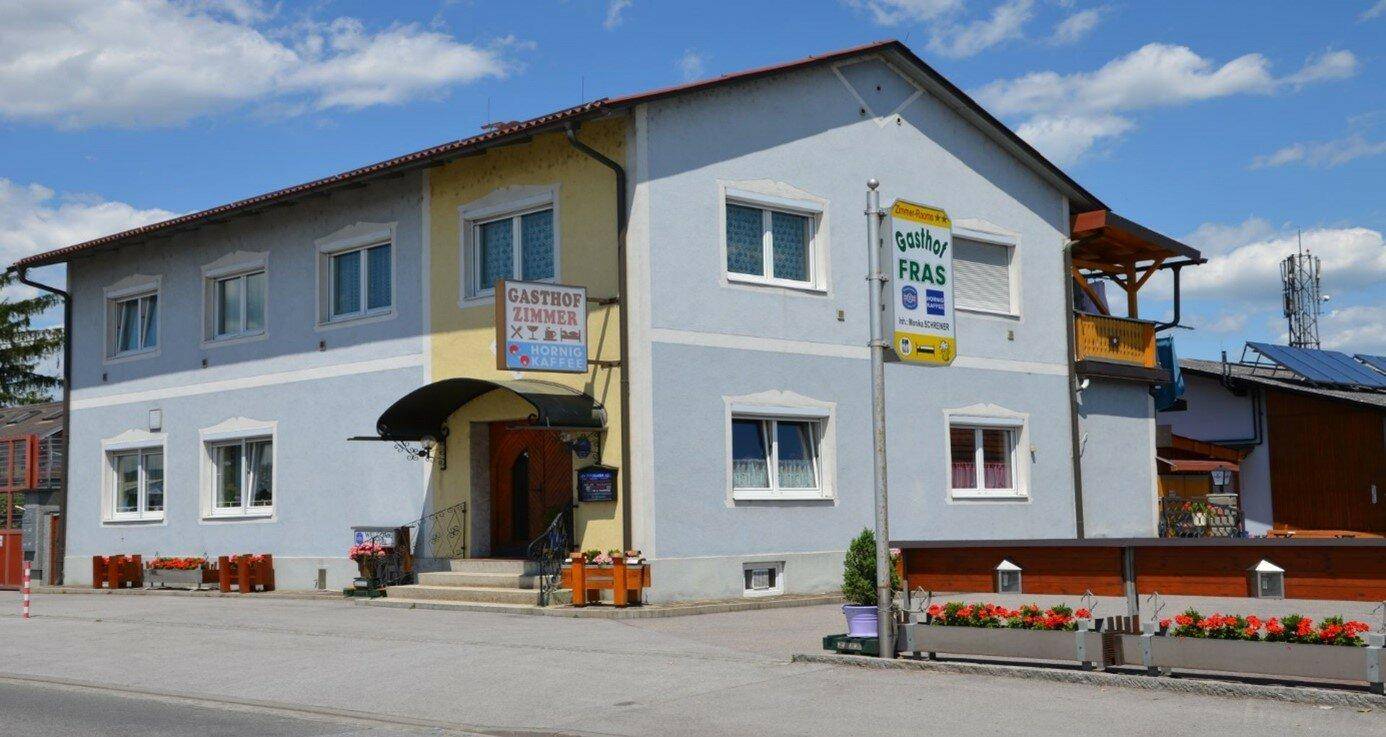 Ansicht des Gasthofs mit hellblauer und gelber Fassade unter einem blauen Himmel.