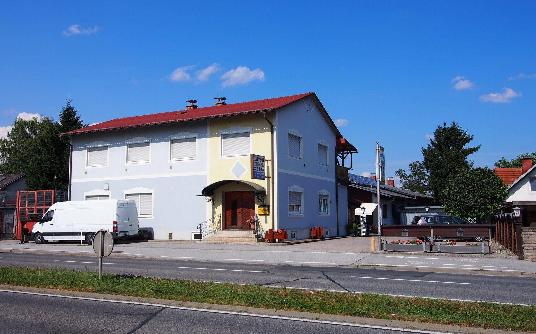 Breitere Außenansicht des Gasthofs mit Parkplatz und Straße an einem sonnigen Tag.
