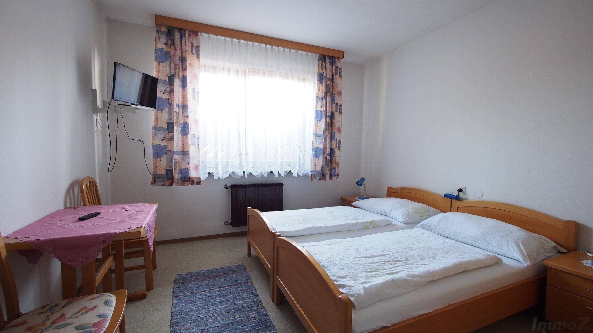 Schlafzimmer mit zwei Einzelbetten, kleinem Tisch und Fernseher an der Wand, Teppichboden.