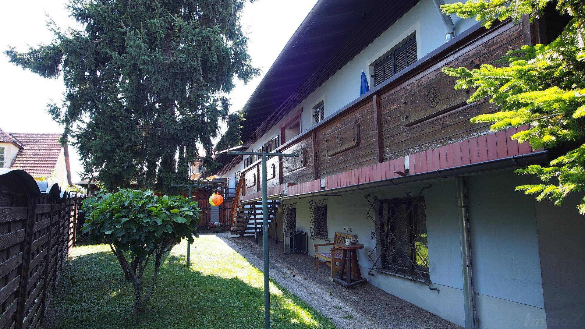 Lange Gebäudeseite mit durchgehendem Balkon, Gartenbereich und hohen Bäumen.