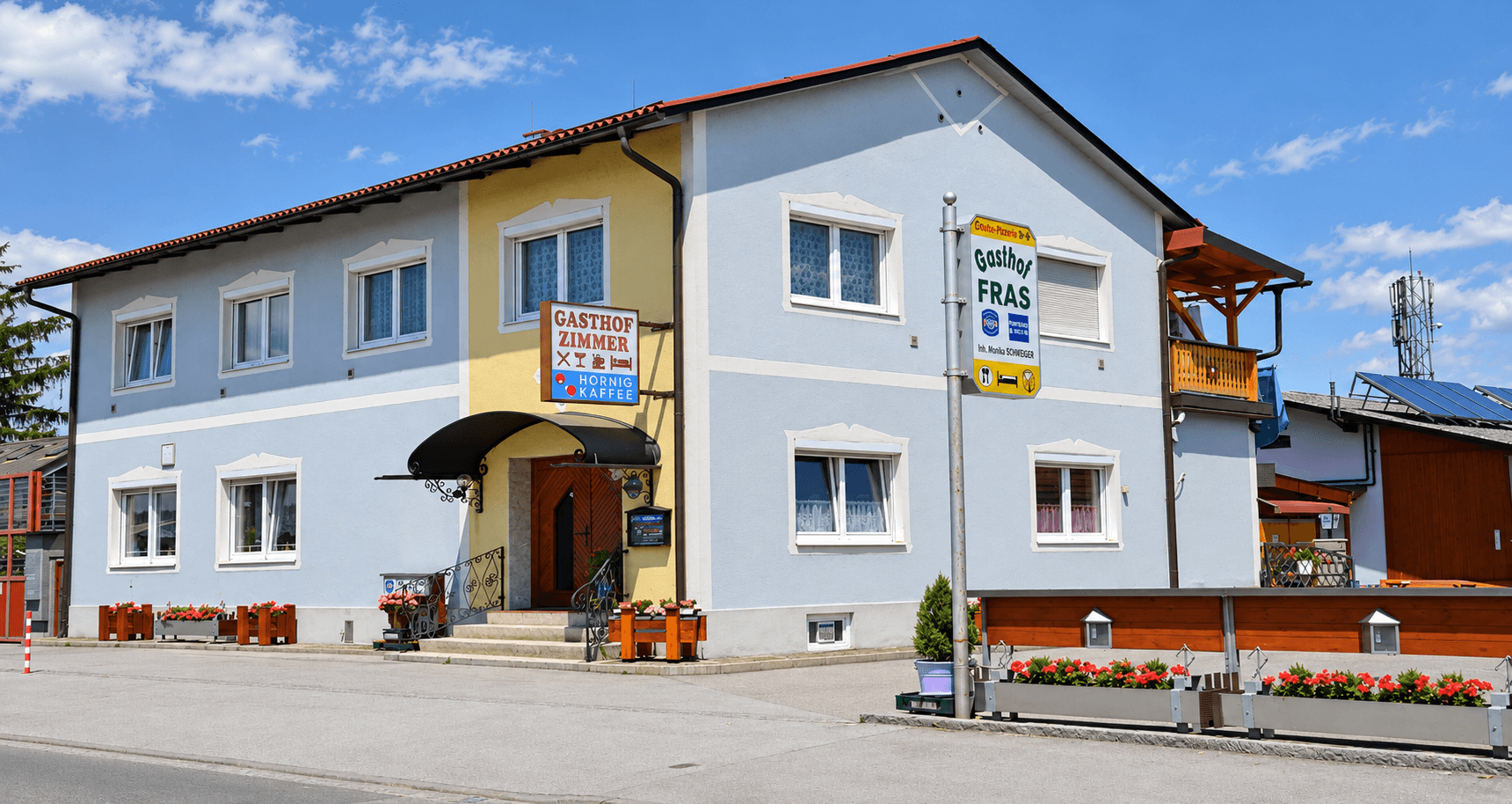 Gasthof mit hellblauer und gelber Fassade, rotem Dach, Balkon und Blumenkästen am Eingangsbereich.