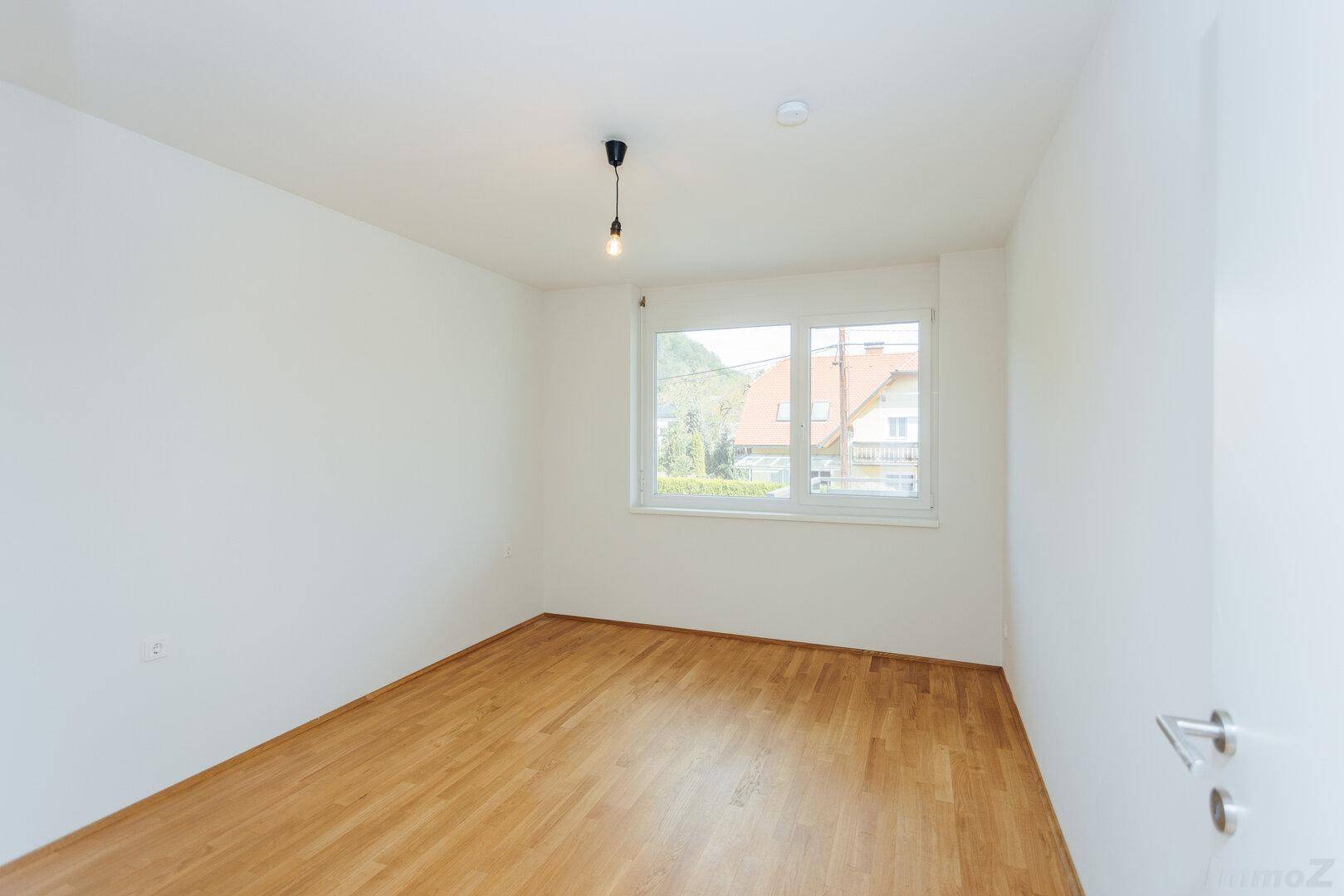 Schlafzimmer mit Parkettboden, großem Fenster und Blick ins Grüne.
