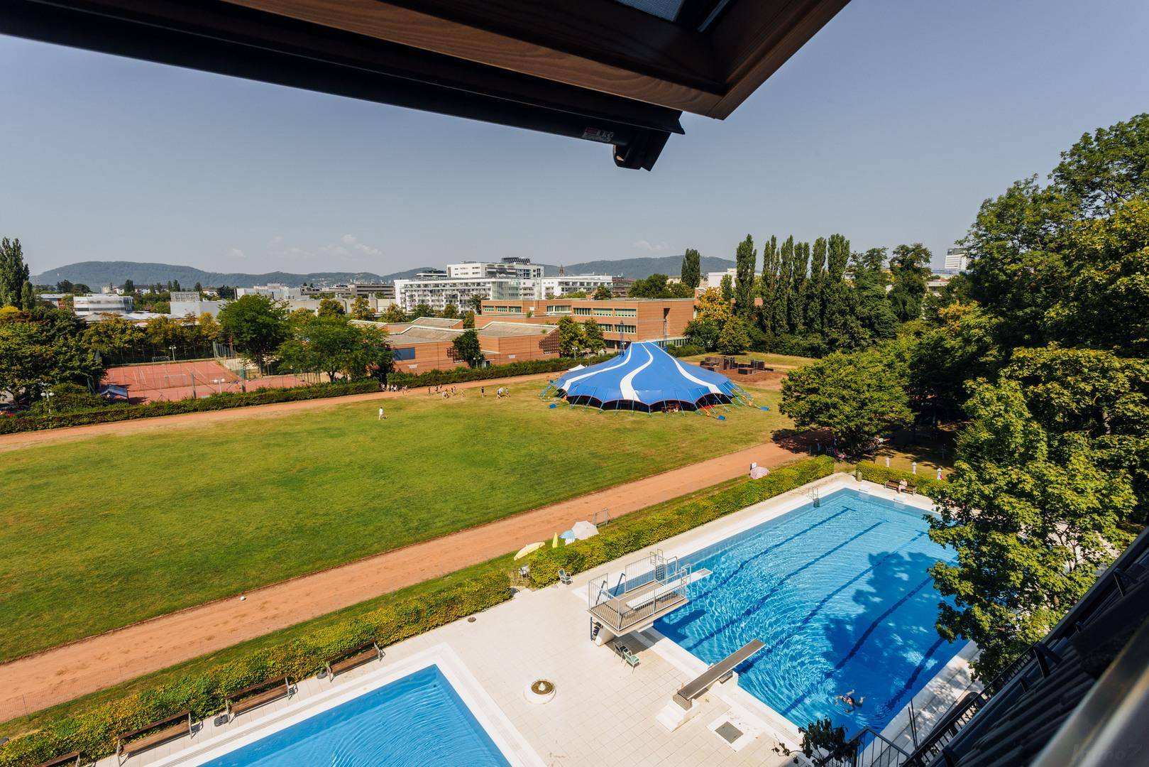 Weitläufiger Blick auf Sportanlagen, Schwimmbad und grüne Umgebung von oben.
