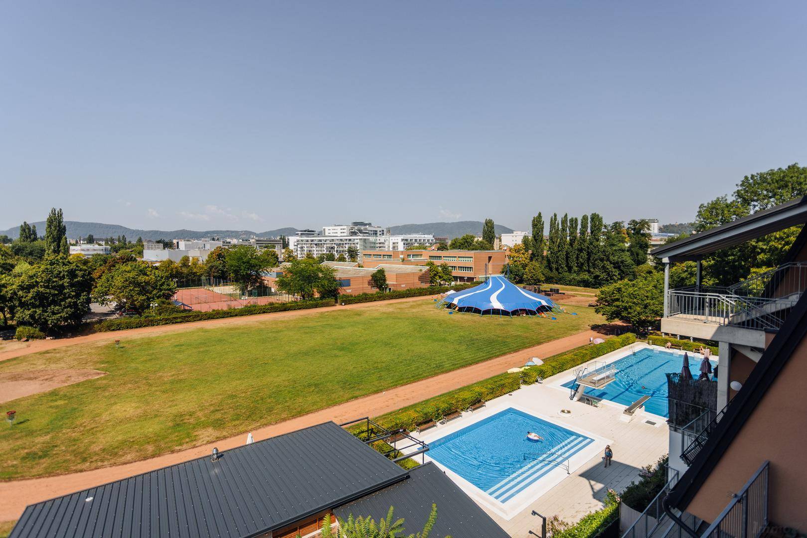 Weitläufiger Ausblick auf eine Sportanlage mit Schwimmbad, Grünflächen und Bergen im Hintergrund.
