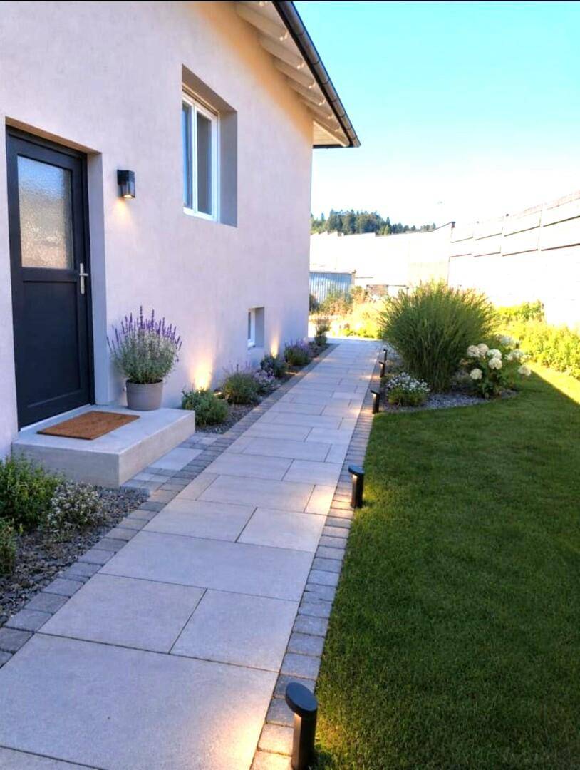Gepflasterter Weg entlang des Hauses mit Rasenfläche, Gartenbeleuchtung und Eingangstür.