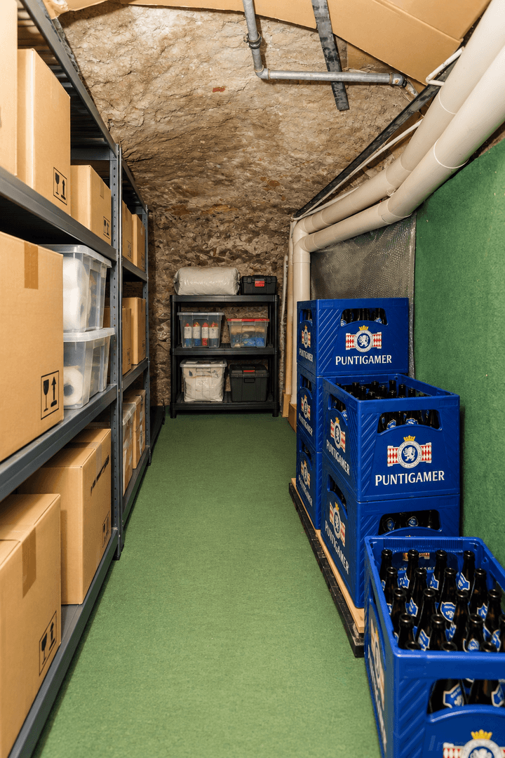 Schmaler Kellergang mit Steinwänden, grünem Teppichboden und Lagerregalen voller Kartons.