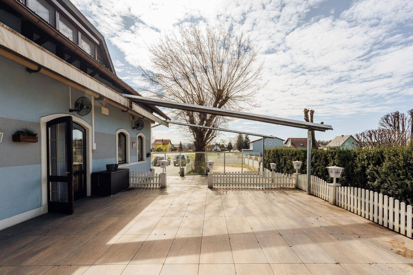 Große gepflasterte Terrasse neben dem Gebäude, umgeben von einem weißen Zaun und einem alten Baum.
